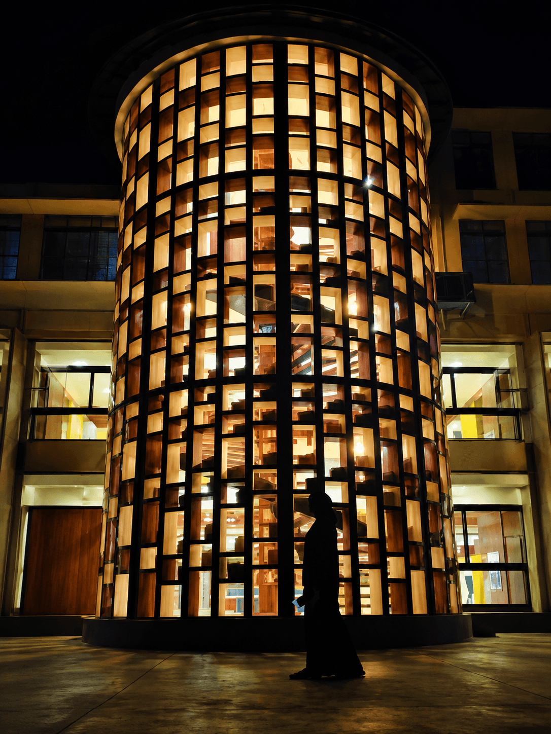 The Devagiri College Library at night: a tall cylindrical reading tower in a black steel grid, lit from within, with a figure walking past at ground level